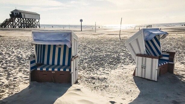 Two beach chairs with striped cushions on a sandy beach, a pier in the background, and a few people in the distance. - Ferienwohnung in St. Peter-Ording (St. Peter-Ording)