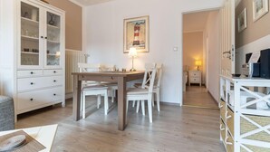 A dining area with a wooden table and chairs, a cabinet with glass doors, a painting of a lighthouse, and a small bedside table with a lamp. - Ferienwohnung in St. Peter-Ording (St. Peter-Ording)