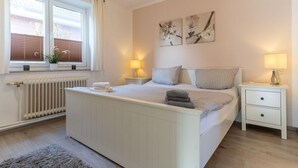 A bedroom with a bed, bedside tables, a radiator, a window with blinds, and artwork on the wall. - Ferienwohnung in St. Peter-Ording (St. Peter-Ording)