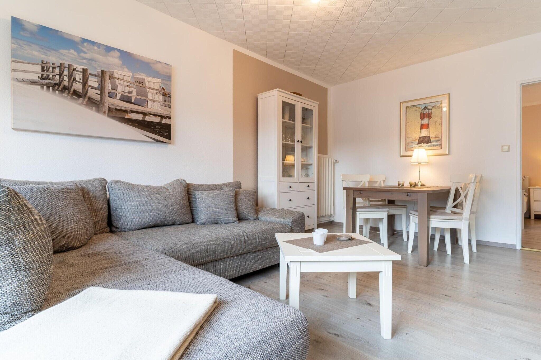 En moderne stue med en grå sofa, et hvidt sofabord, et spiseområde med bord og stole, samt et skab med glasdøre.