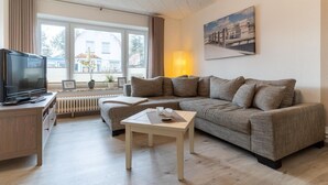 A living room with a sofa, a coffee table, a TV, and a window with curtains. - Ferienwohnung in St. Peter-Ording (St. Peter-Ording)