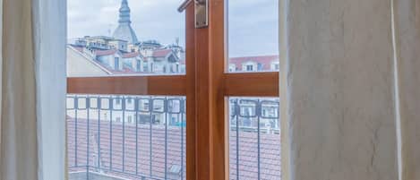 A view from a window with a wooden frame and a metal latch, overlooking a cityscape with buildings and a tall tower.