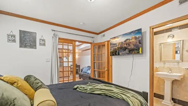 A bedroom with a bed, pillows, a painting, a bathroom with a sink, and a view of the city.