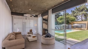 Una sala de estar moderna con sofá, mesa de centro y un otoman redondo. Hay una puerta de vidrio que da a una zona de piscina.