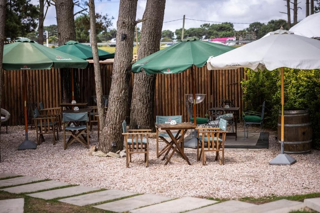 An outdoor seating area with wooden tables and chairs, covered by green and white umbrellas, and surrounded by trees and a wooden fence.
