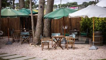 An outdoor seating area with wooden tables and chairs, covered by green and white umbrellas, and surrounded by trees and a wooden fence.