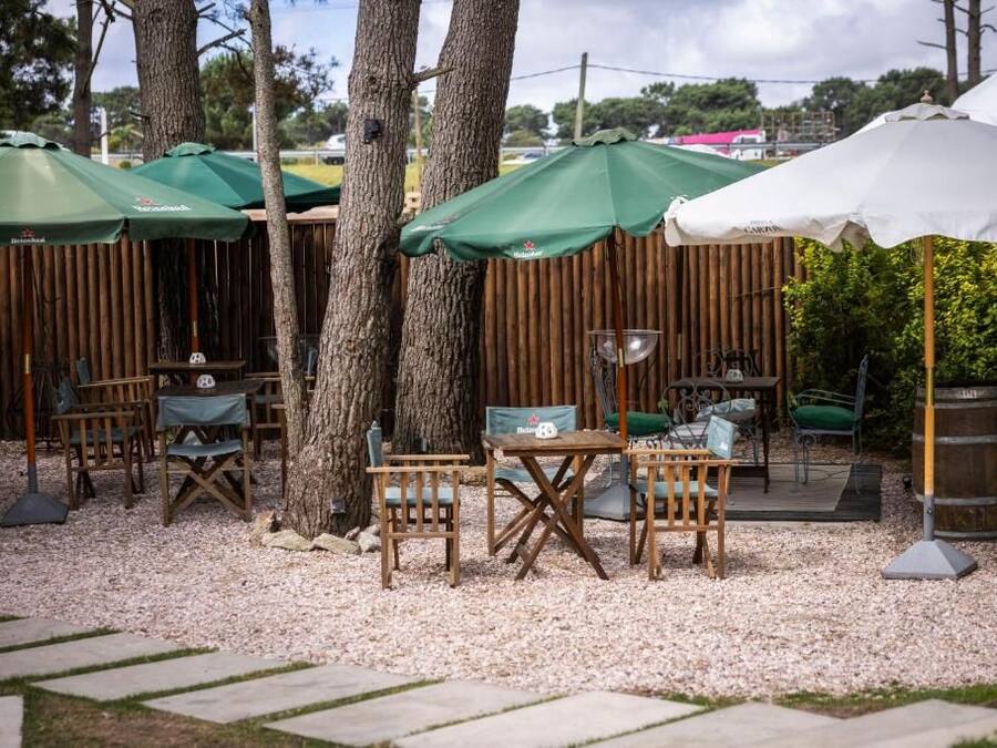 An outdoor seating area with wooden tables and chairs, covered by green and white umbrellas, and surrounded by trees and a wooden fence.