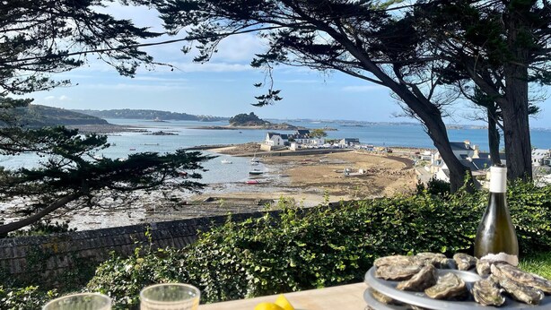 A table with oysters, a bottle of wine, and glasses, overlooking a coastal area with boats and buildings. - Holiday Home in Terenez with Sea Views (Plougasnou)