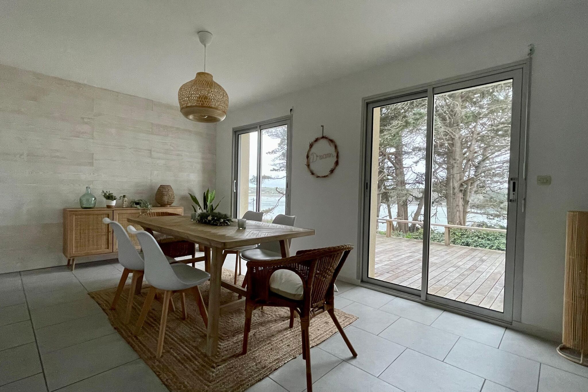 Un coin repas avec une table en bois, des chaises blanches, un luminaire suspendu en fibres tressées et une vue sur une terrasse extérieure, accessible par de grandes portes-fenêtres coulissantes.