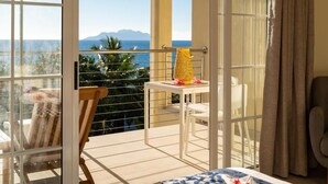 Un balcon avec une table et des chaises, un fauteuil avec un coussin à rayures, et une vue sur la mer et les montagnes.