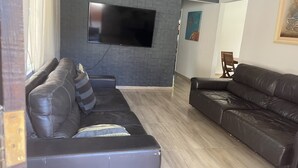 Una sala de estar moderna con dos sofás de cuero, un televisor de pantalla plana montado en una pared con diseño de ladrillo, y piso de madera.