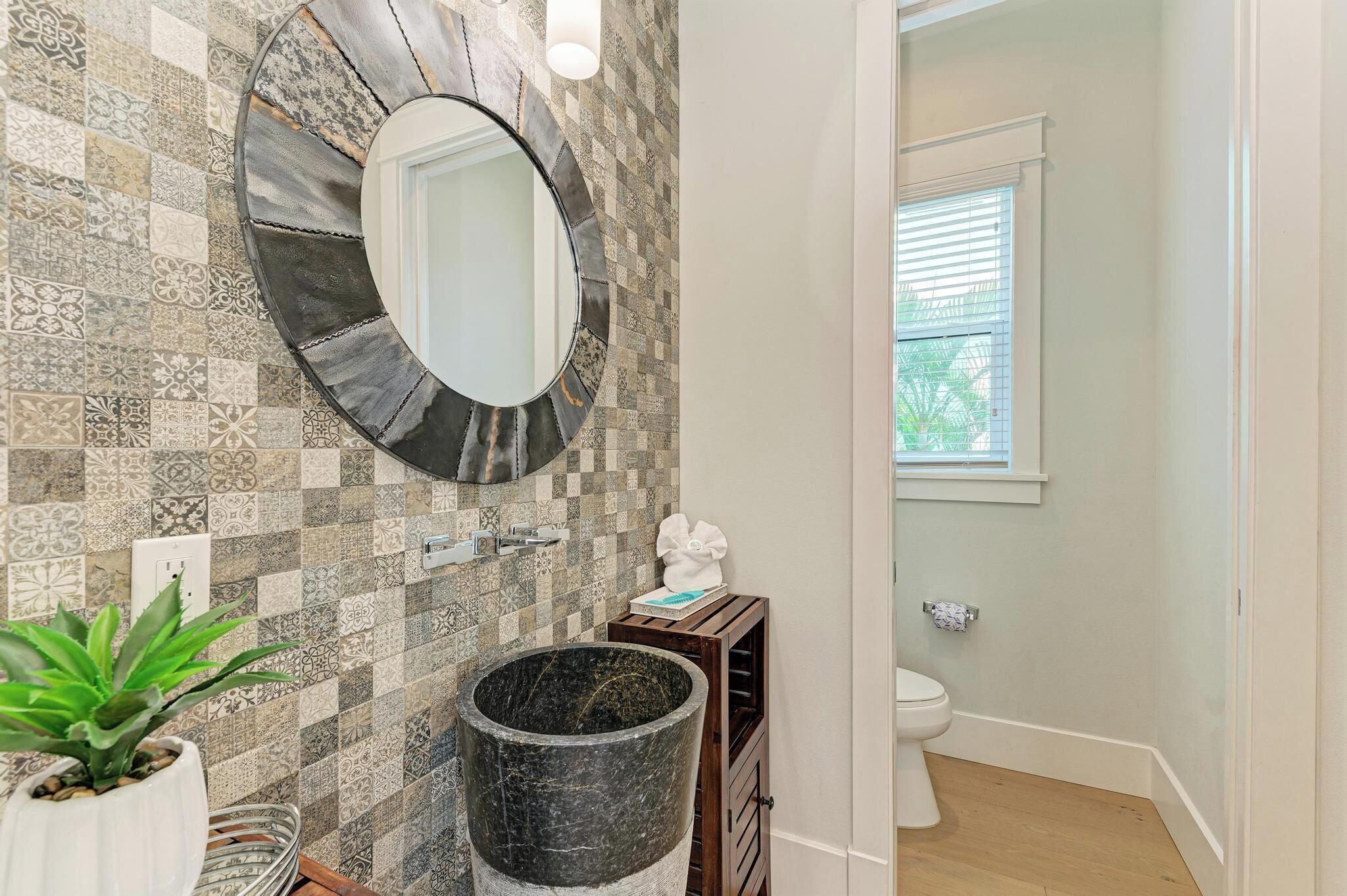 A bathroom with a mosaic tile wall, a round mirror, a sink, a toilet, and a potted plant.