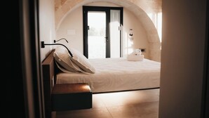Ein Schlafzimmer mit einem Bett, einem Nachttisch, einer Lampe, einem Fenster und einer gewölbten Steinwand.