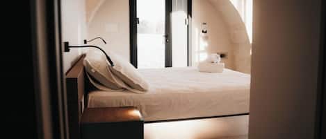 A bedroom with a bed, a nightstand, a lamp, a window, and an arched stone wall.
