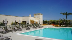 Ein Poolbereich mit Liegestühlen und Sonnenschirmen, ein Gebäude mit flachem Dach und einem strahlend blauen Himmel.