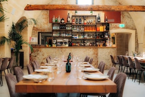 Un restaurant avec une longue table en bois dressée pour le repas, un bar avec des étagères remplies de bouteilles et un mur en pierre rustique.
