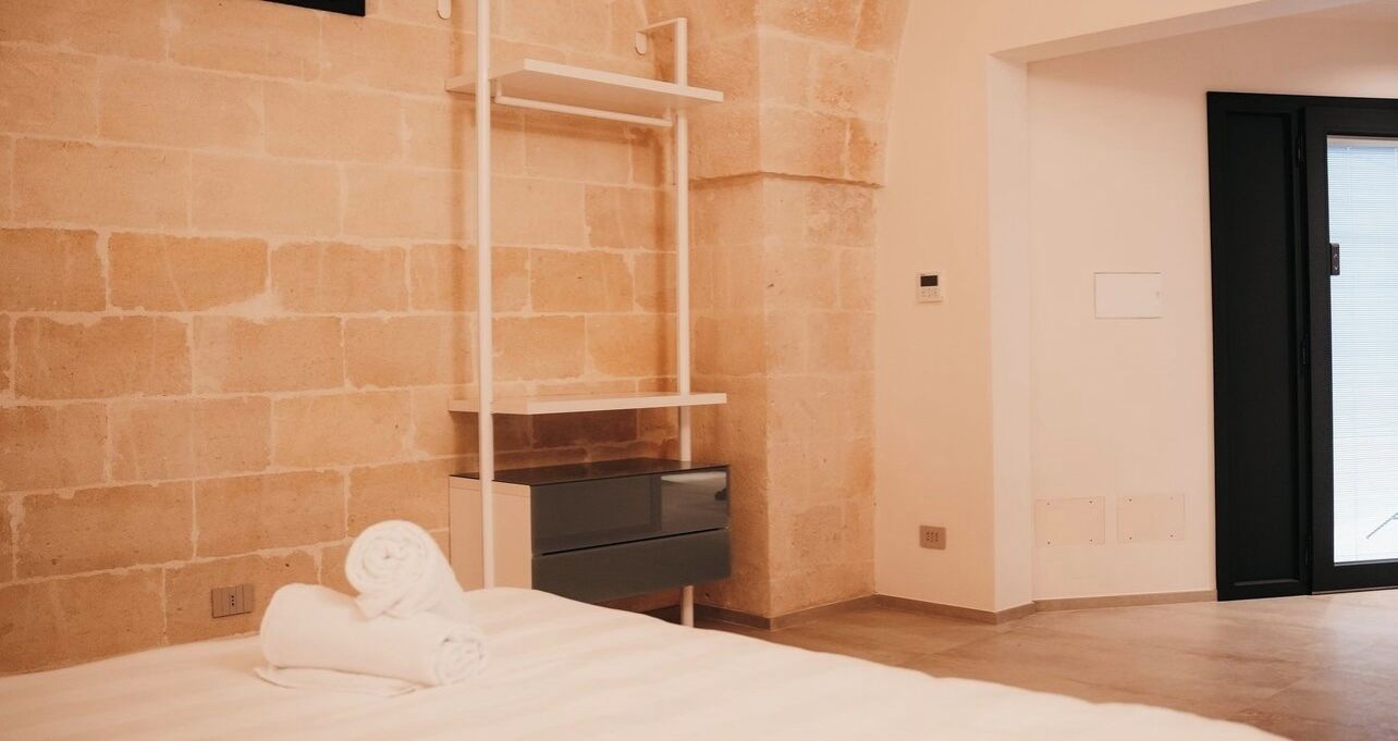 A bedroom with a bed, a white shelf unit, a dark-colored dresser, and a stone wall.