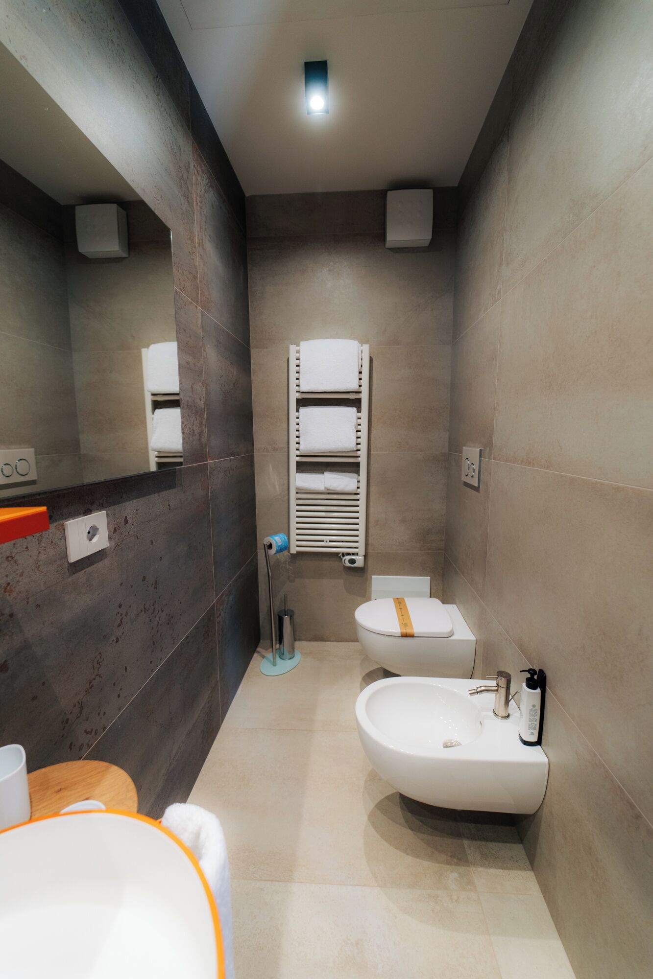 Un baño moderno con inodoro, bidé y lavamanos. Hay un perchero con toallas, un espejo y una lámpara en el techo.