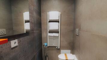 Un baño moderno con inodoro, bidé y lavamanos. Hay un perchero con toallas, un espejo y una lámpara en el techo.