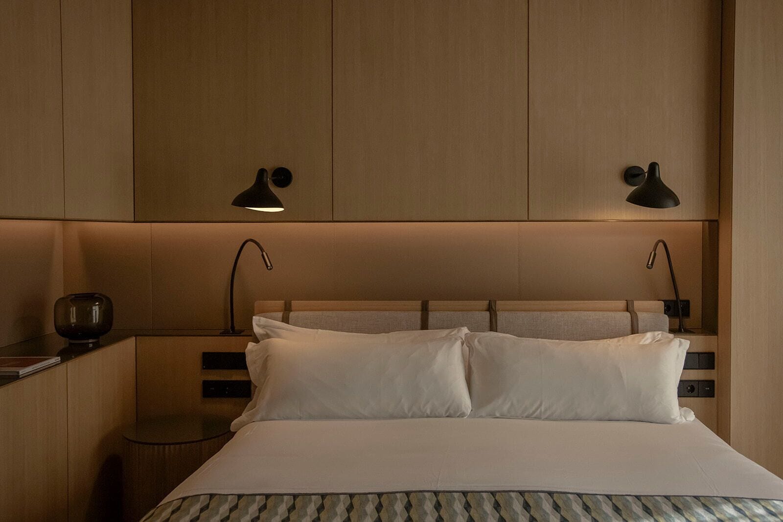 Ein modernes Hotelzimmer mit einem großen Bett, zwei Nachttischlampen, einem Nachttisch mit Lampe und einem kleinen Beistelltisch mit einem dekorativen Gegenstand.