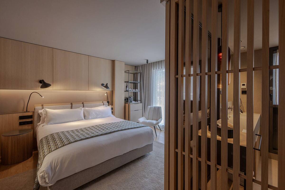Une chambre d’hôtel moderne avec un grand lit, des tables de chevet, une armoire et une salle de bain visible à travers une cloison en bois.