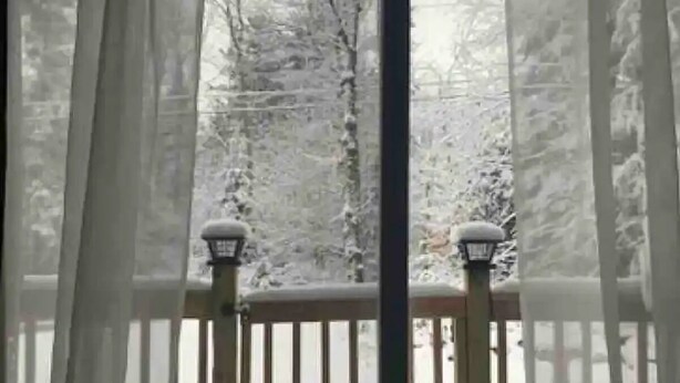 Vista a través de una ventana con cortinas translúcidas, mostrando un paisaje nevado con una terraza de madera y dos faroles.