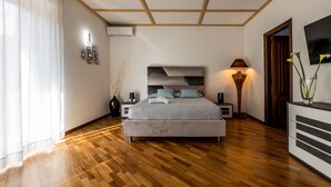 A modern bedroom with a wooden floor, a bed with a grey headboard, bedside tables, a lamp, and a TV on the wall. - I Barcaioli (Giardini Naxos)