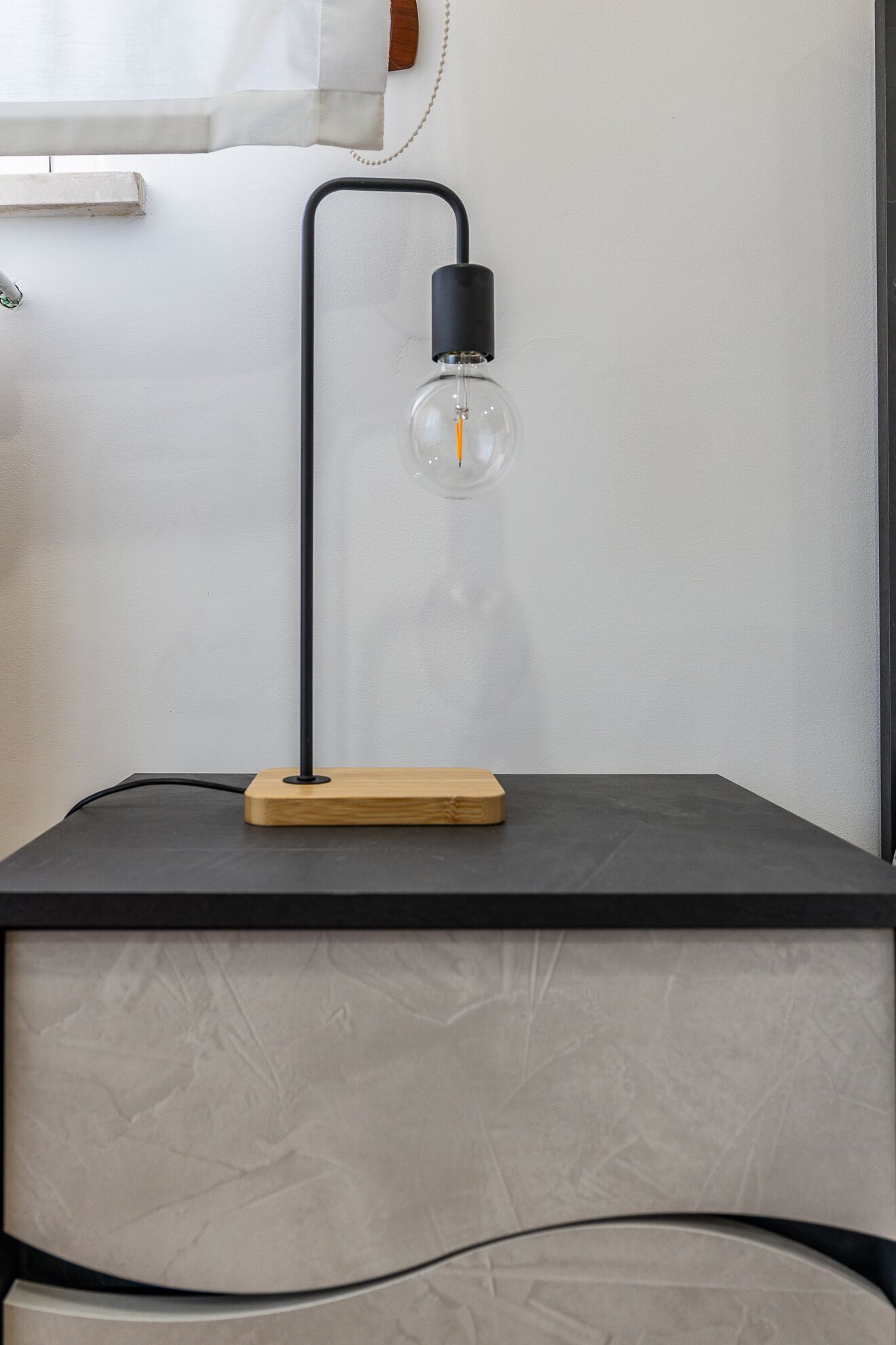 Una lámpara de escritorio moderna con pantalla de vidrio transparente y base de madera, sobre una mesa oscura frente a una pared con textura.