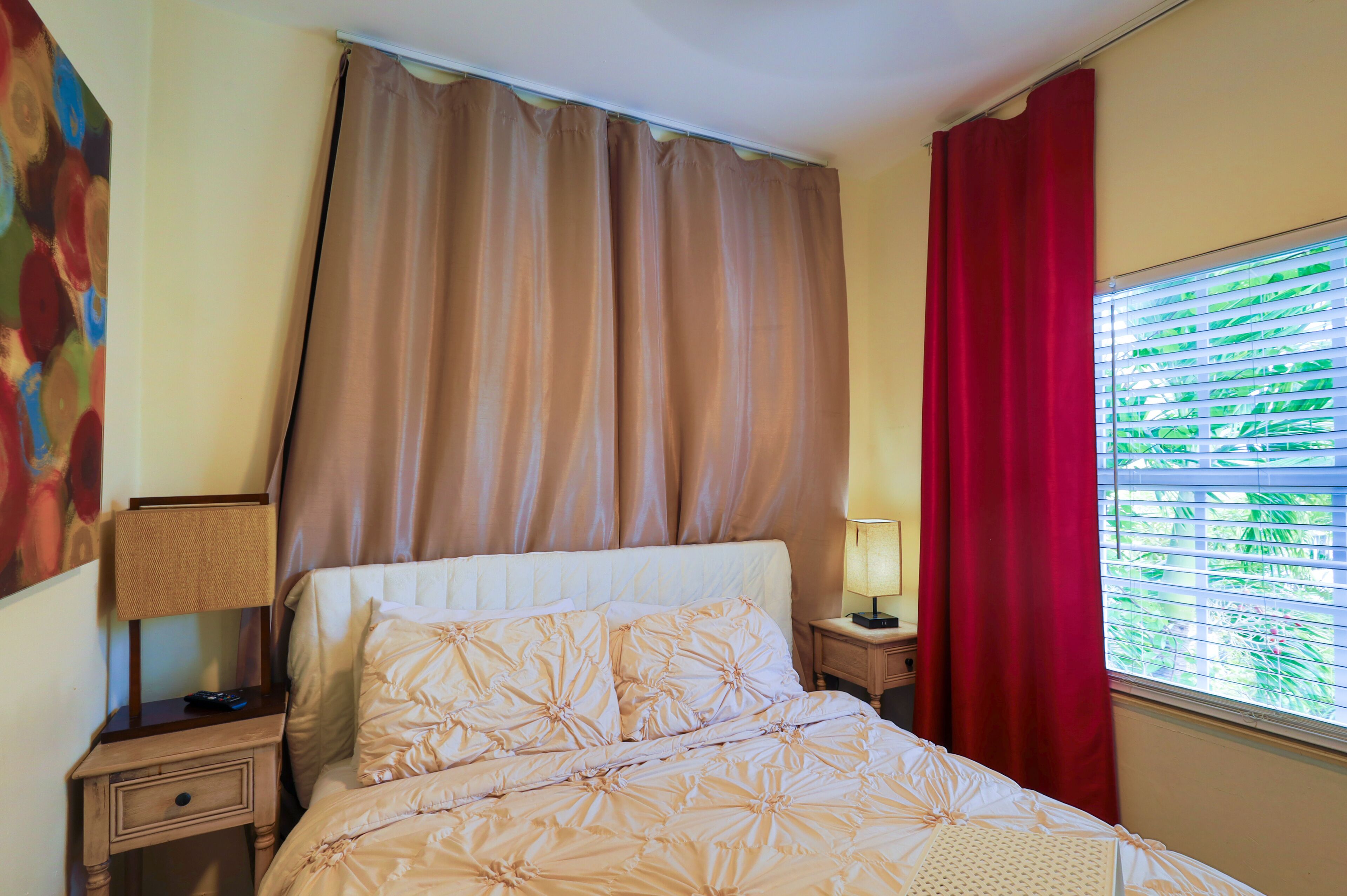 Une chambre d’hôtel avec un lit, une table de chevet, une lampe, une fenêtre avec des stores et un rideau rouge.