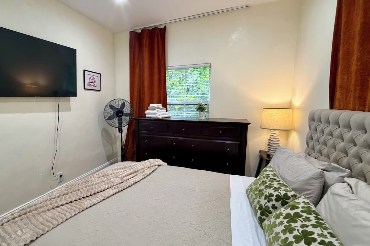 Een slaapkamer met een bed, een aan de muur bevestigde televisie, een dressoir, een ventilator en een raam met gordijnen.