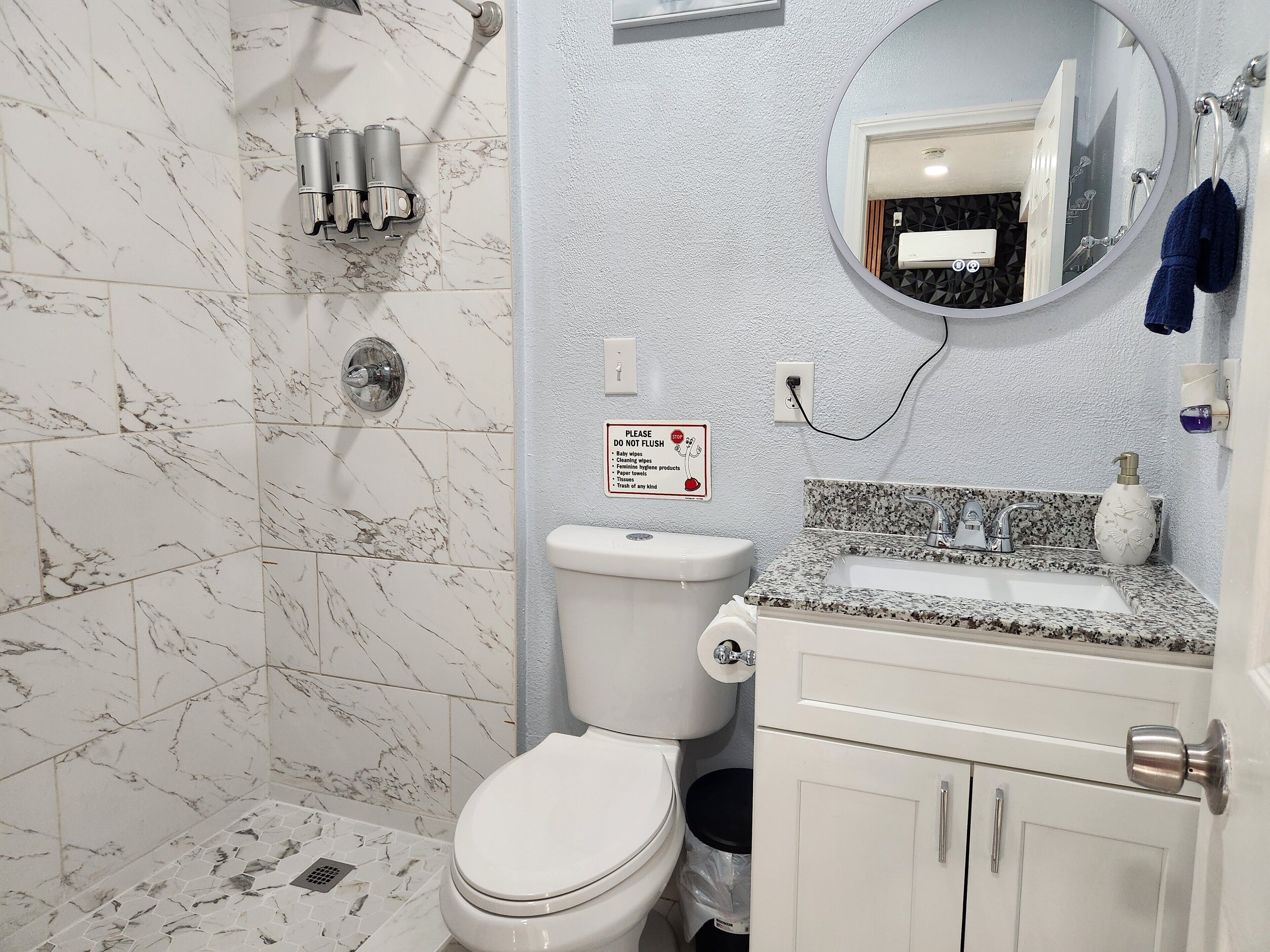 Ein Badezimmer mit Toilette, Waschbecken und Dusche. Ein Schild ist an der Wand angebracht, und über dem Waschbecken befindet sich ein Spiegel.