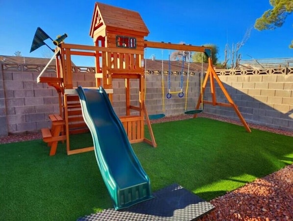 A wooden play structure with a slide and swings, surrounded by a brick wall and green artificial turf.