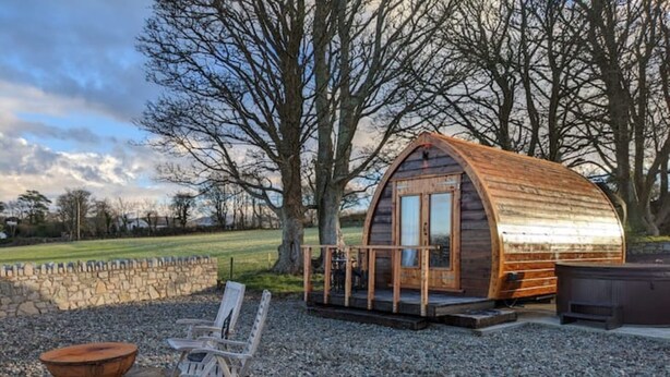A rustic wooden cabin with a curved roof, a stone wall, a fire pit, and a wooden chair.