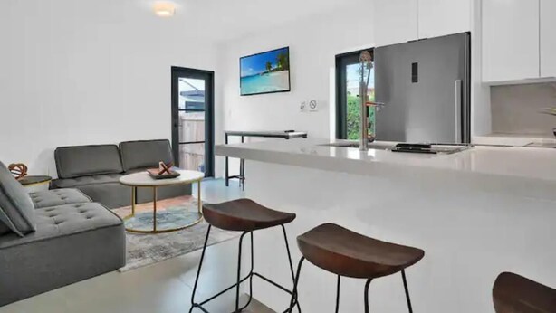 A modern kitchen with white cabinets, a stainless steel refrigerator, and a dining area with a round table and bar stools. - Villa 3 in Miami (Miami)