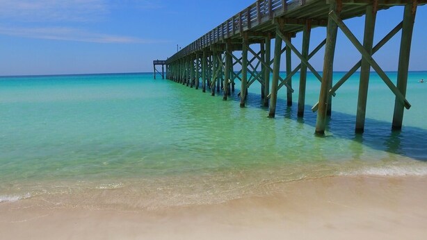 Un muelle de madera que se extiende hacia aguas turquesas cristalinas, con una playa de arena al final.