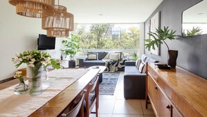 A modern living room with a dining area, a sofa set, and a large window offering a view of greenery.