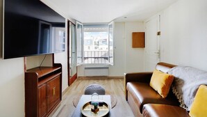 A living room with a flat-screen TV, a brown leather sofa, a coffee table, and a window with a view of buildings. - Paris Marais Best WE MERRIS (Paris)
