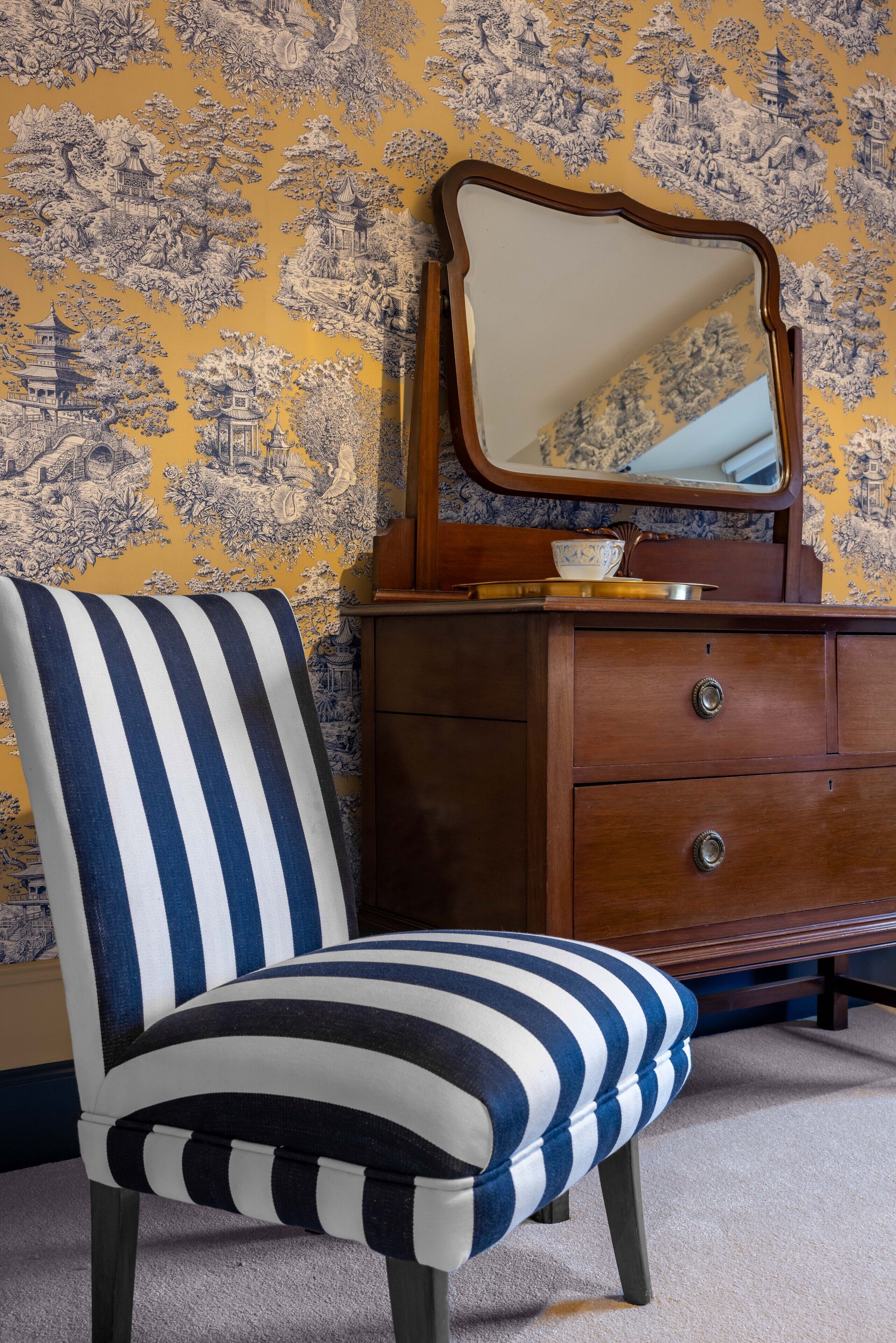 A striped chair with a wooden dresser and a mirror in a room with patterned wallpaper.