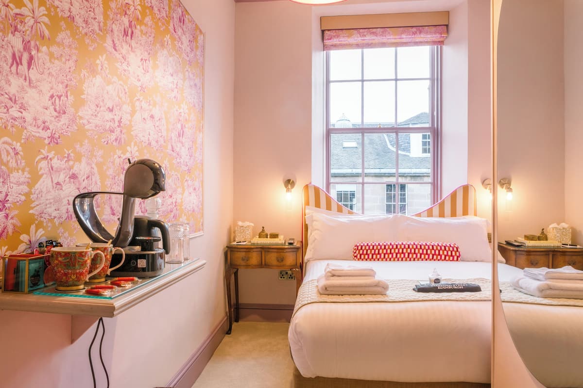 Hotelkamer met een bed, bureau met koffiezetapparaat, spiegel en raam met uitzicht op gebouwen.