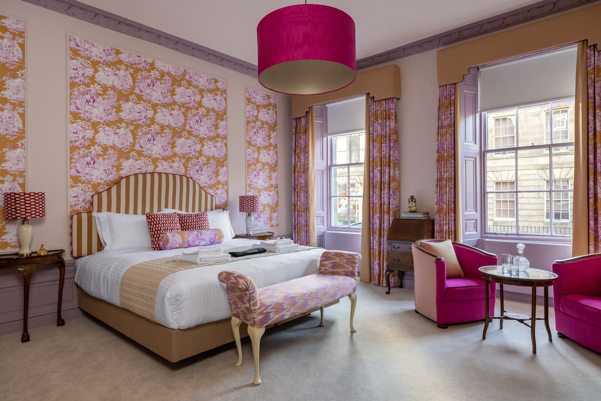 Een slaapkamer met een groot bed, een bankje, een stoel, een tafel en een raam met gordijnen.
