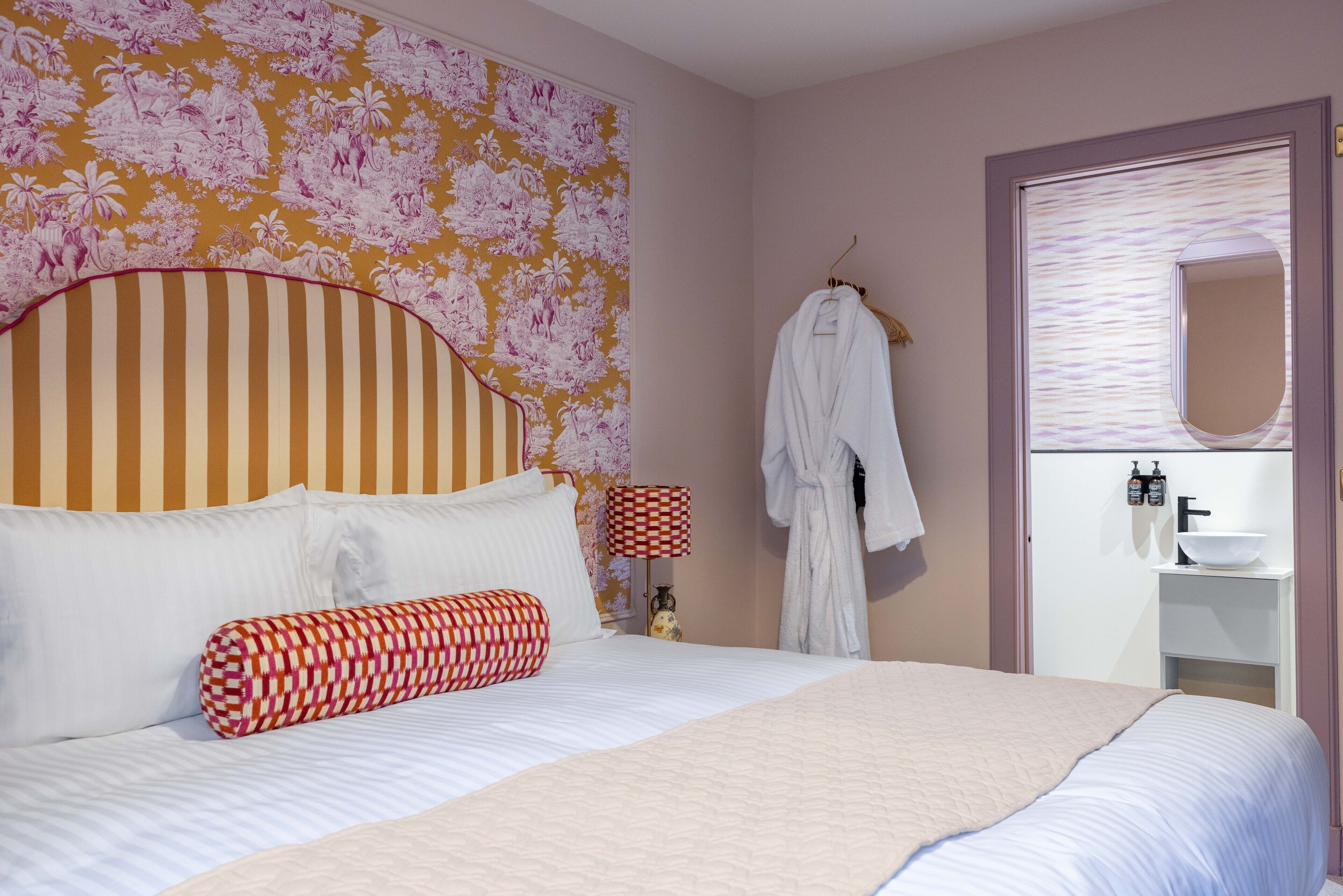 A hotel room with a large bed, striped headboard, floral wallpaper, a hanging robe, and a bathroom with a sink and mirror.
