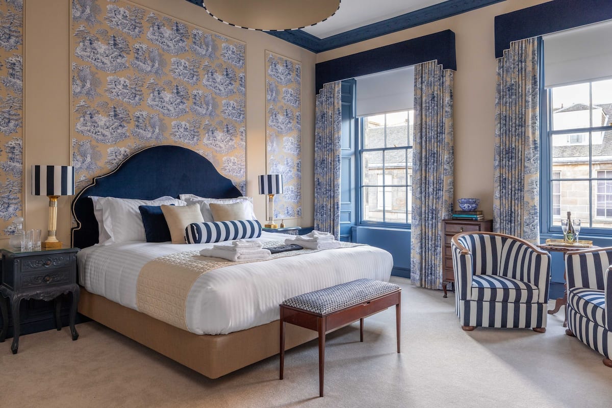 Een slaapkamer met een groot bed, een nachtkastje, een bankje, gestreepte fauteuils en uitzicht op buiten.