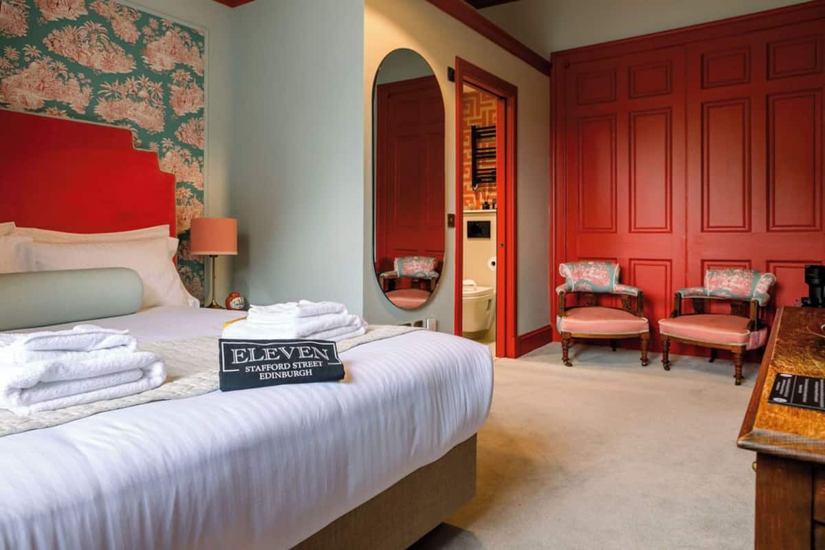 Een hotelkamer met een net opgemaakt bed, een rode deur en twee fauteuils.
