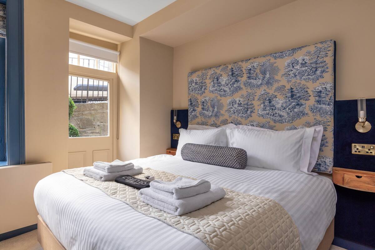 Een net opgemaakt bed met witte lakens en een gewatteerde deken, een hoofdbord met een blauw-wit patroon en een nachtkastje met een lamp.