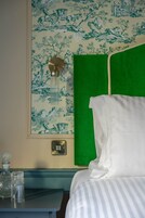 A neatly made bed with a green headboard, a framed floral wallpaper, a bedside table with a lamp, and a wall socket.