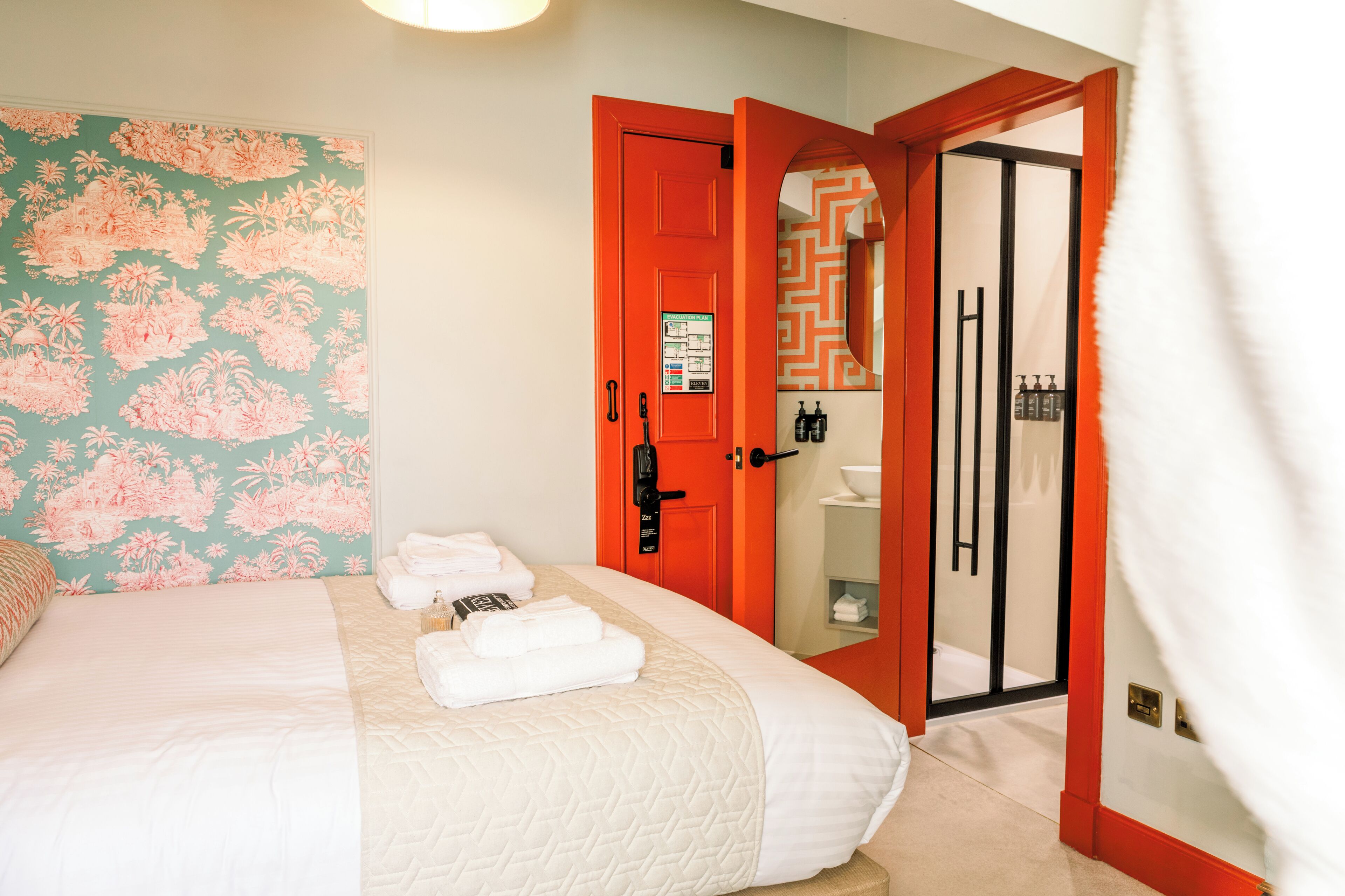 Een hotelkamer met een bed, een badkamer met douche, en een deur naar een gang.