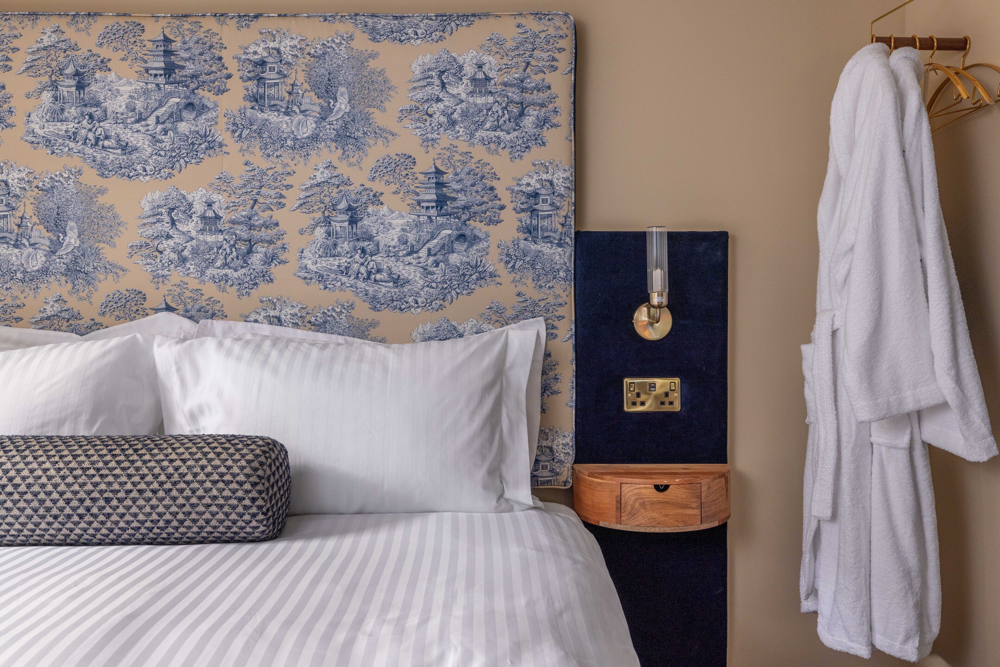 Un lit bien fait, avec une literie blanche et rayée, un coussin à motifs, une décoration murale bleue et blanche, une table de chevet en bois avec un tiroir bleu, et une robe blanche accrochée à un crochet.