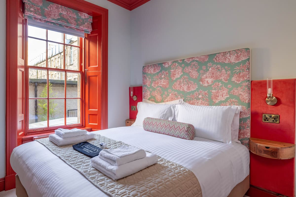 Een net opgemaakt bed met gevouwen handdoeken en een sprei, een raam met rode randen en een behang met een bloemendesign aan de muur.