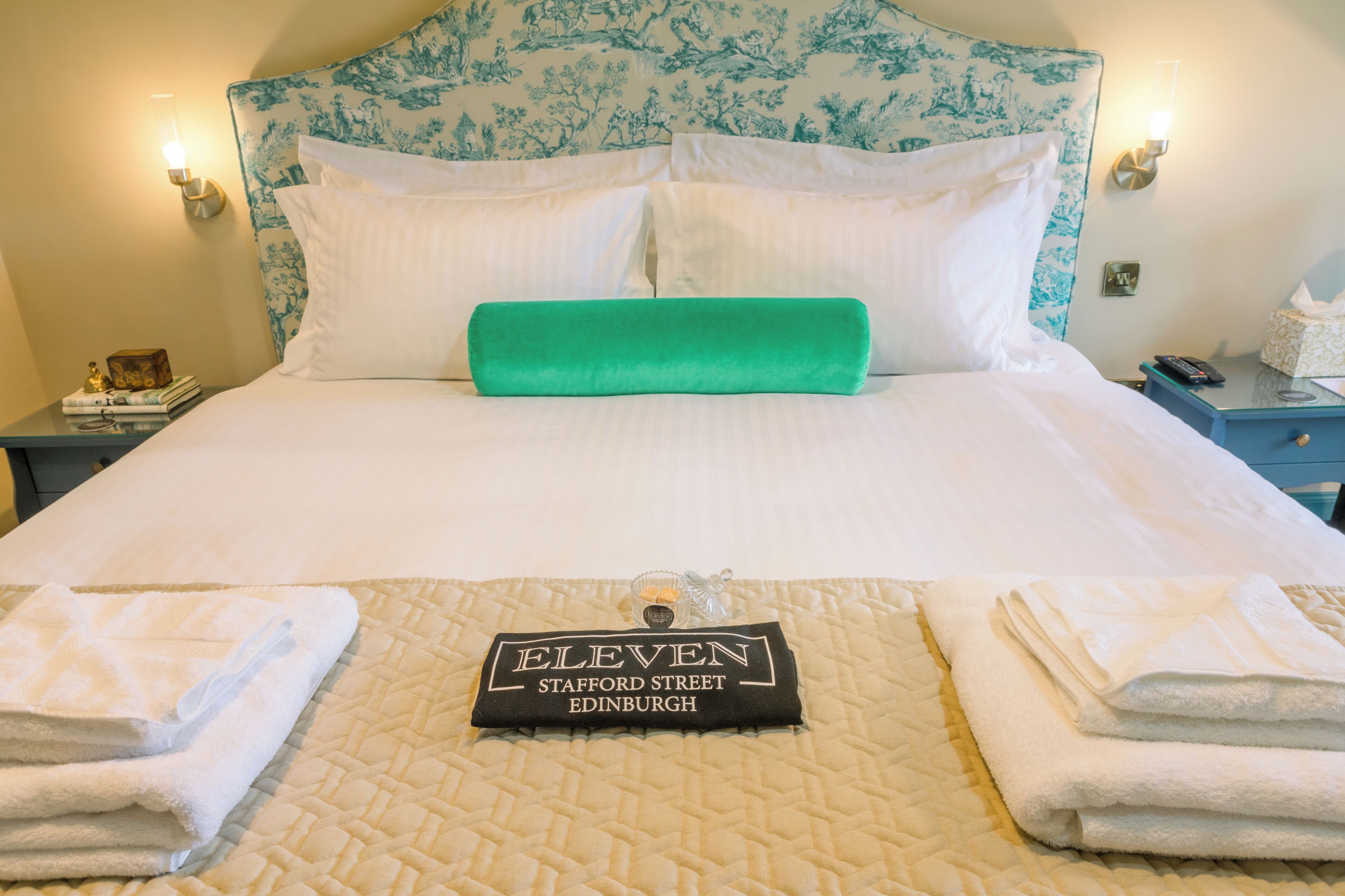 A neatly made bed with a quilted headboard, a green accent pillow, and a sign indicating the hotel name and location.