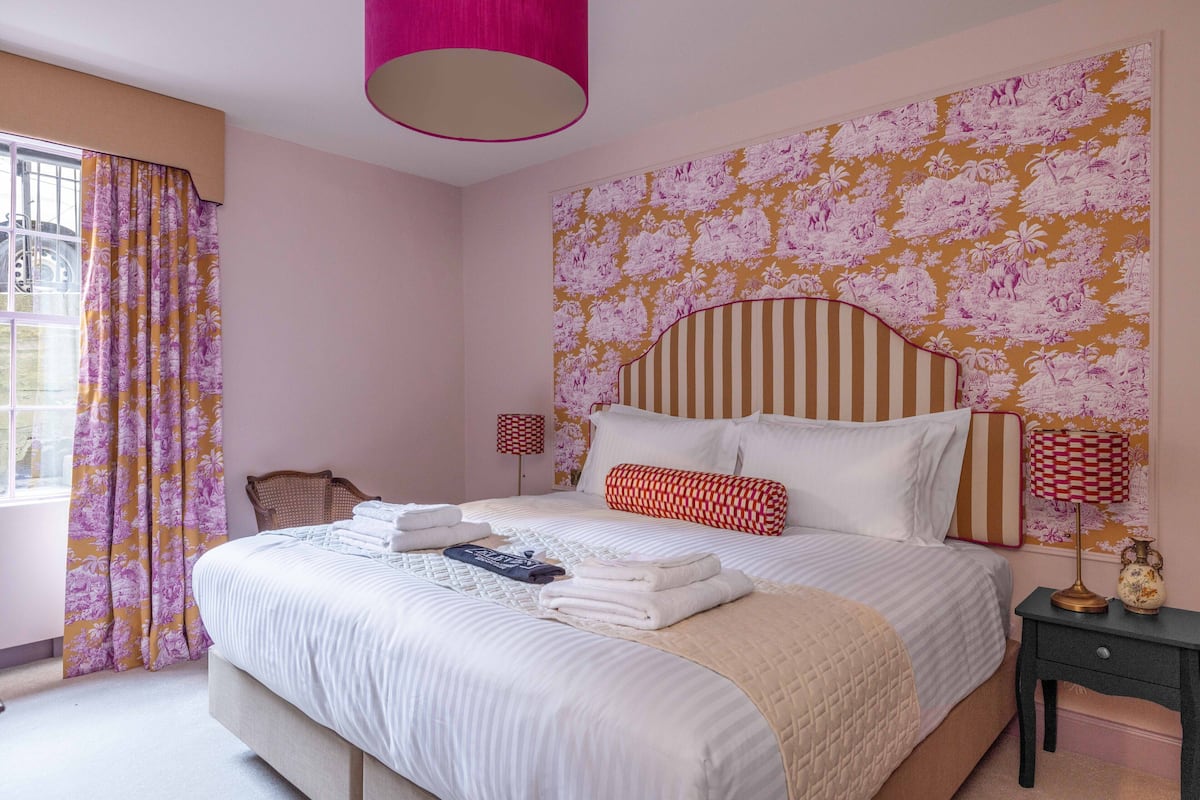 Een net opgemaakt bed met witte lakens, een gestreept hoofdbord en een wand met bloemenprint.